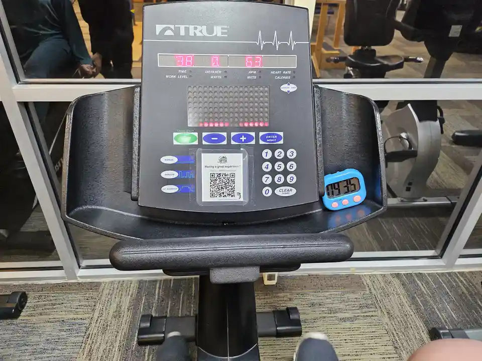 A close-up of a True brand exercise bike console showing workout metrics like time (38), RPM (63), and heart rate. A small…