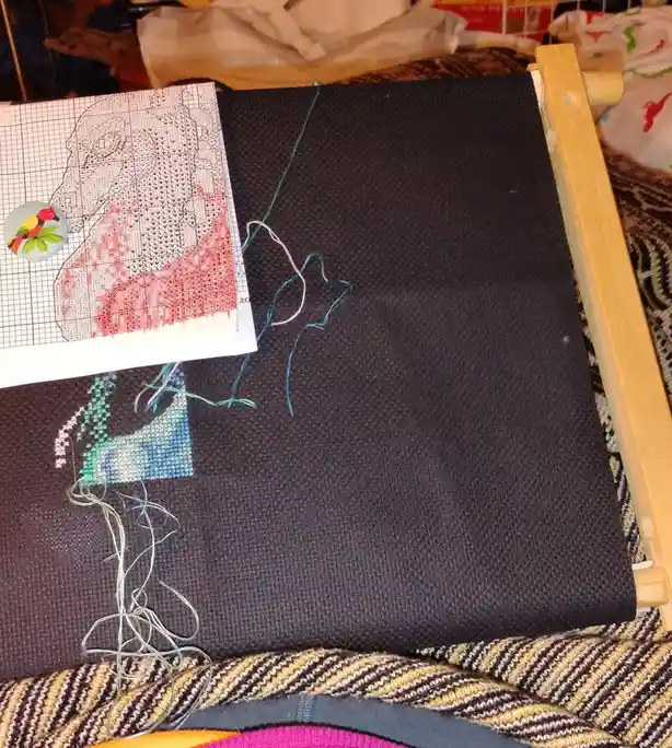 A close-up photo shows a cross-stitch project in progress on dark fabric stretched on a wooden frame. A partially completed…