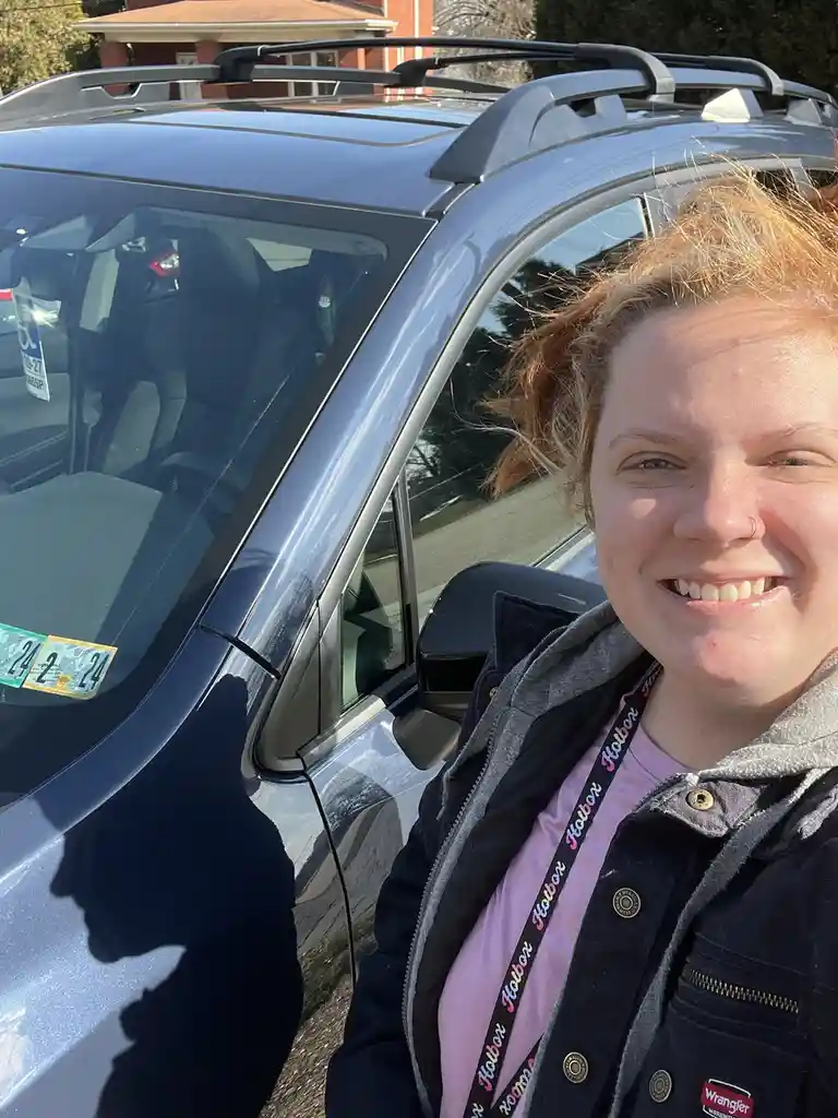A smiling person with reddish hair stands beside a silver SUV, taking a selfie. The car has a roof rack and visible 2024…