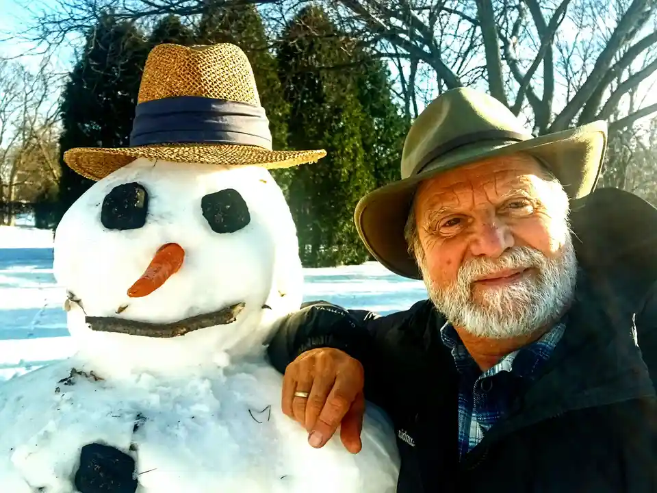 A man with a white beard and a wide-brimmed hat stands beside a snowman in a snowy outdoor setting, smiling at the camera…