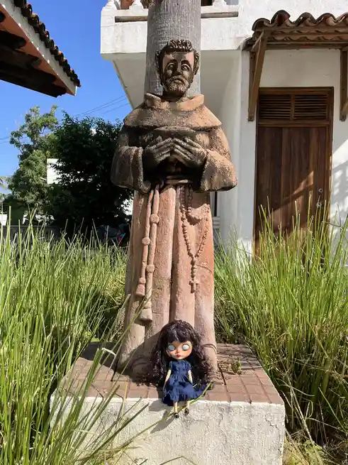A small doll with large blue eyes and dark curly hair sits at the base of a weathered stone statue of a robed monk or saint…