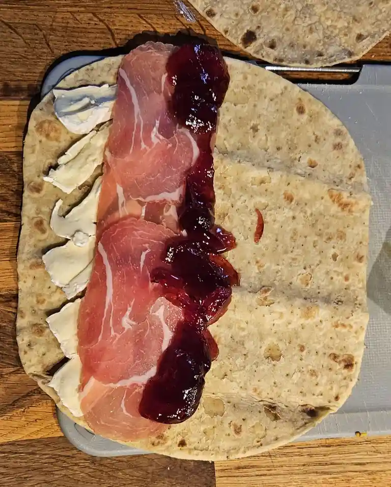 A flatbread tortilla is laid out with a line of creamy white cheese, a line of pink cured meat, and a line of dark red jam…