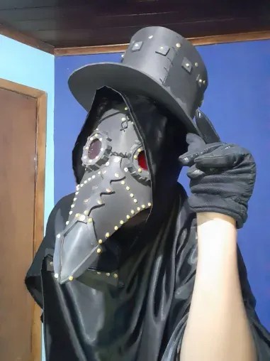A person wearing a black plague doctor costume with a long beak mask, studded hat, and gloves, pointing at the camera. The…