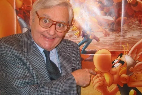 An older man with glasses and a checkered suit points at a cartoon character on a poster behind him. The poster features a…