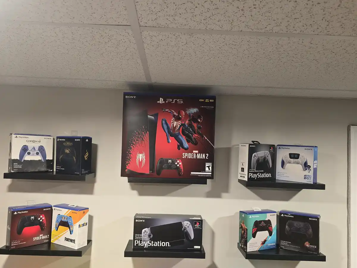 A wall of black shelves displays multiple Sony PlayStation product boxes, including a large Spider-Man 2 PS5 console box…