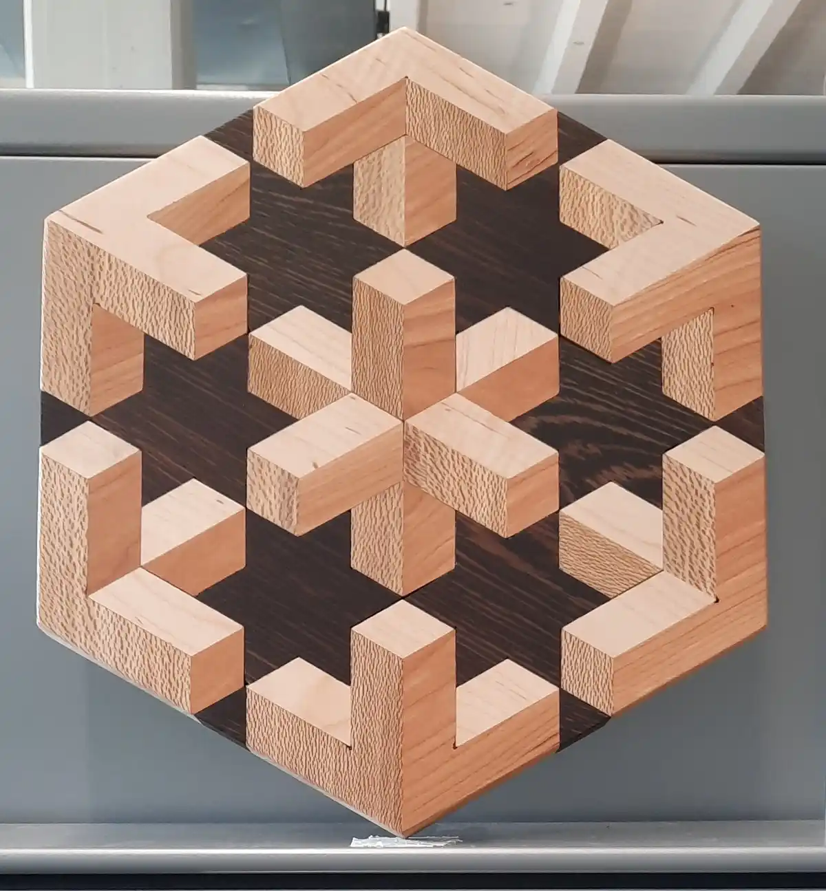 A wooden 3D puzzle with light and dark wood pieces arranged in a geometric, interlocking pattern resembling a cube or…