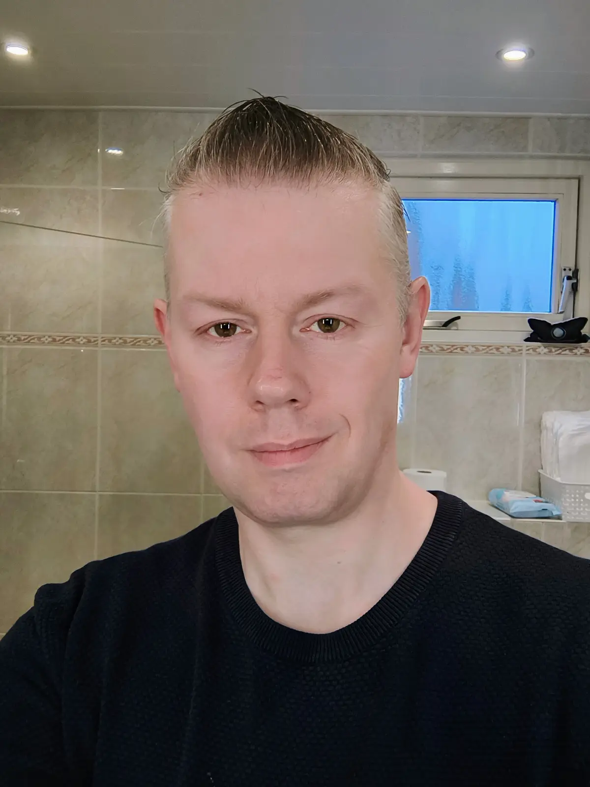 A man with wet, slicked-back hair and a subtle smirk stares at the camera in a bathroom with tiled walls and a frosted…