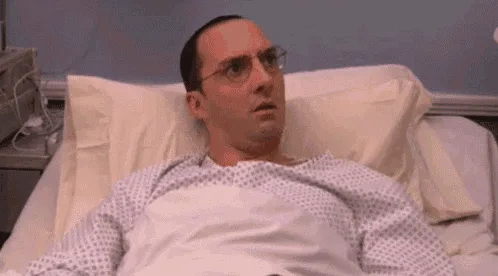 A man in a hospital gown and glasses lies in a hospital bed, initially looking shocked or alarmed, then transitioning to a…