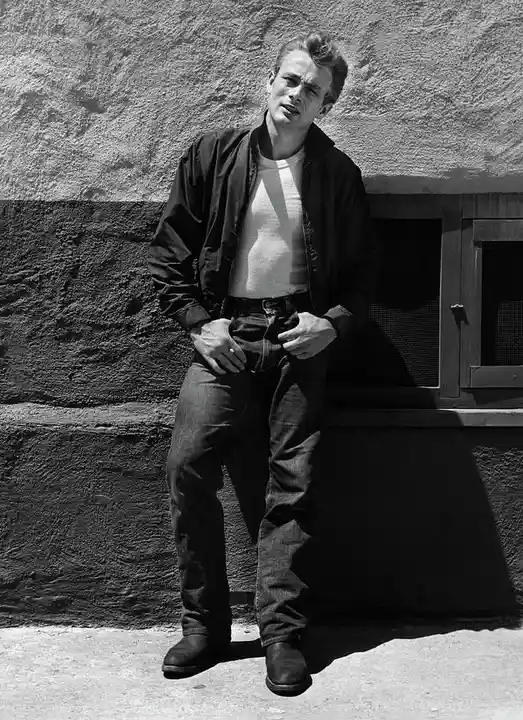 a black and white photo of a young man in a bomber jacket and jeans leaning against a textured wall, hands on his belt, with…