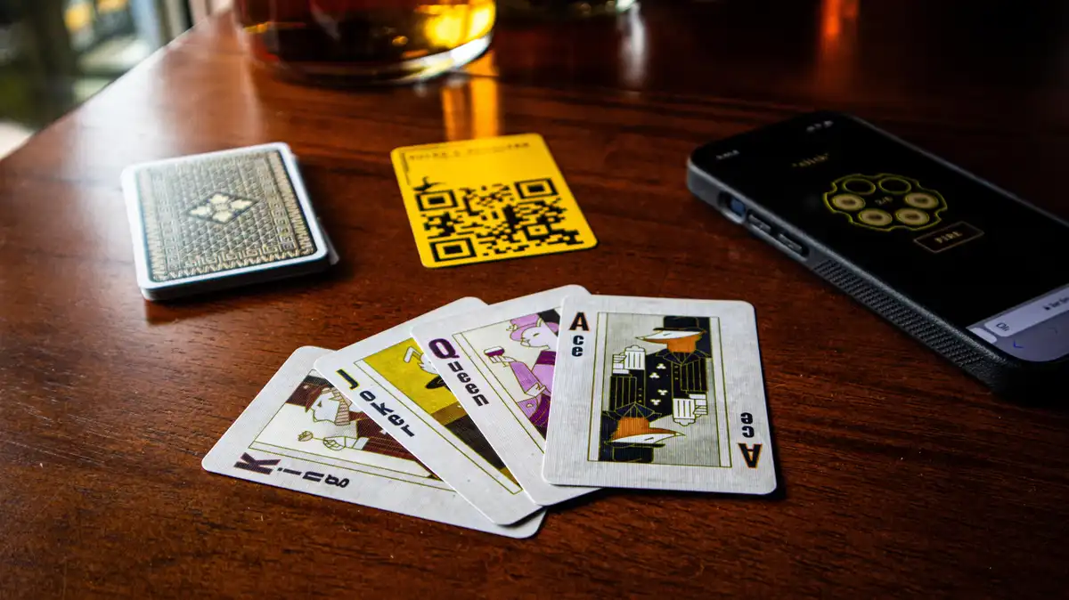 A wooden table holds a spread of playing cards showing an ace, queen, jack, and king, alongside a smartphone displaying a…