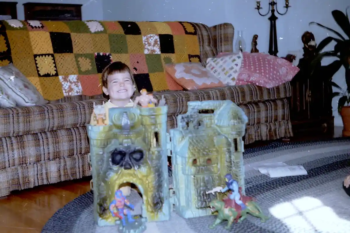 A smiling young boy sits behind two large, green plastic He-Man castle playsets on a patterned rug in a living room. The…