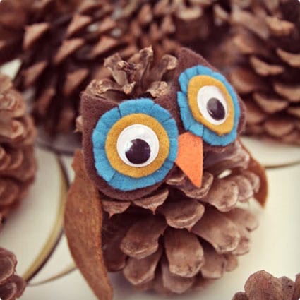 A close-up photo of a pinecone decorated with felt to look like an owl, featuring large blue and yellow felt eyes with black…
