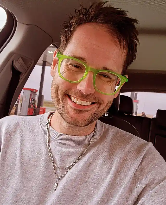 a man with brown hair and a beard is smiling while wearing bright green-framed glasses inside a car, with a gas station…