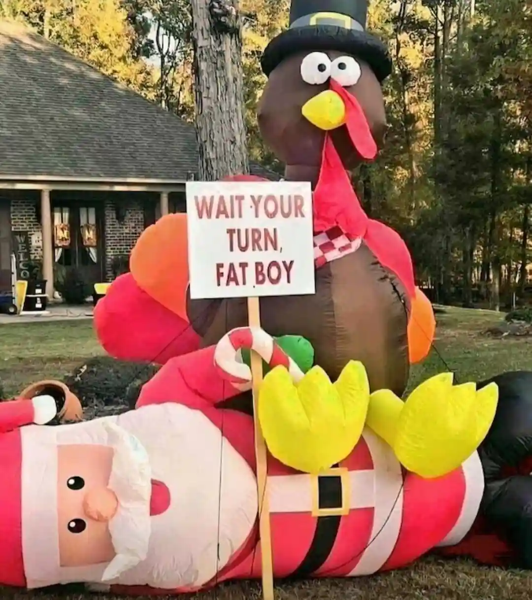 An inflatable turkey wearing a pilgrim hat holds a sign that reads 'WAIT YOUR TURN, FAT BOY' while standing over a fallen…