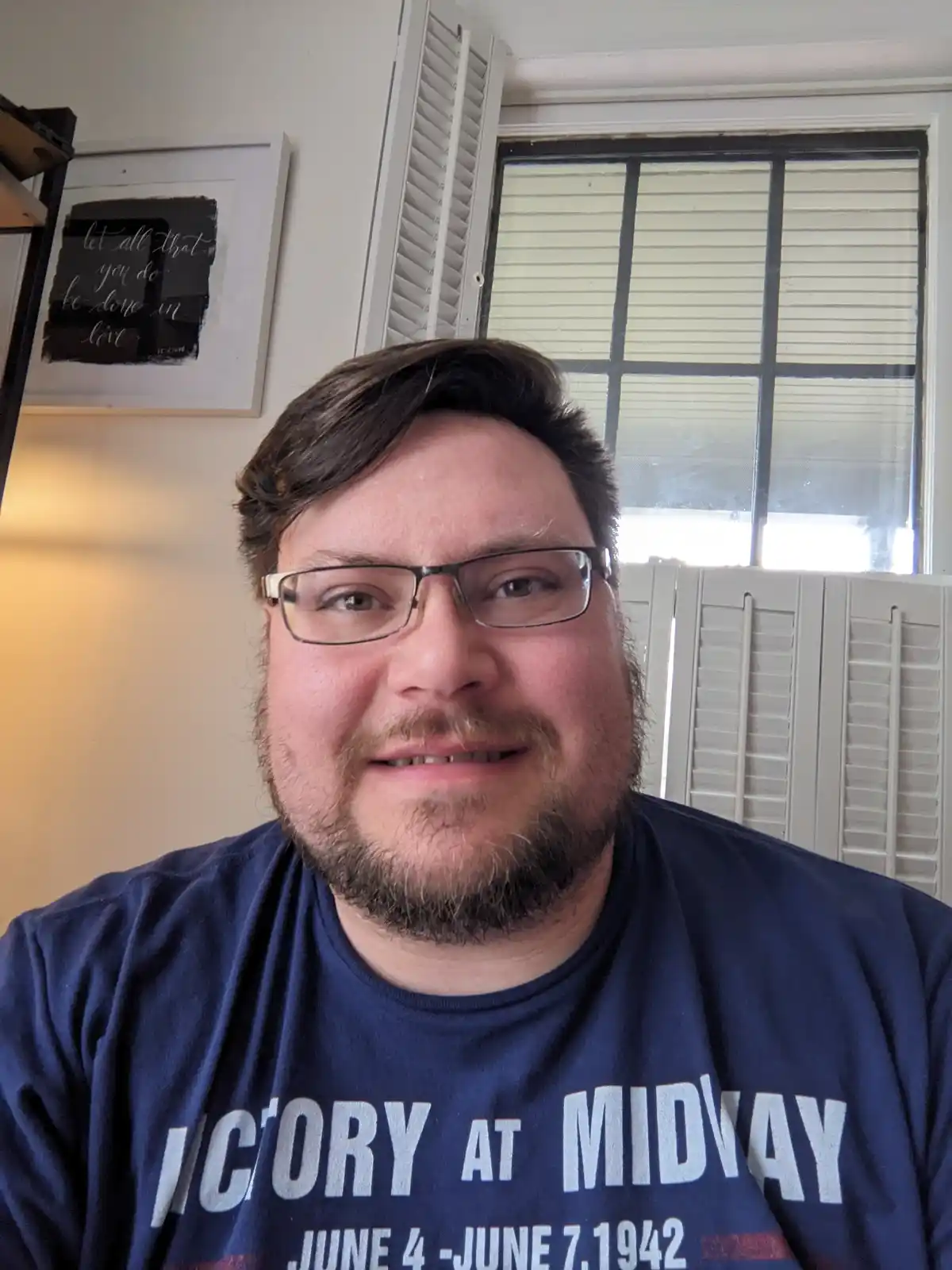 A man with glasses and a beard smiles at the camera while wearing a navy blue t-shirt that reads 'VICTORY AT MIDWAY JUNE 4…