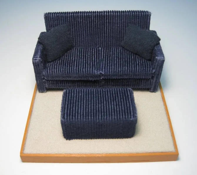a miniature, dark blue corduroy couch with two matching pillows and a matching ottoman, displayed on a beige carpeted…
