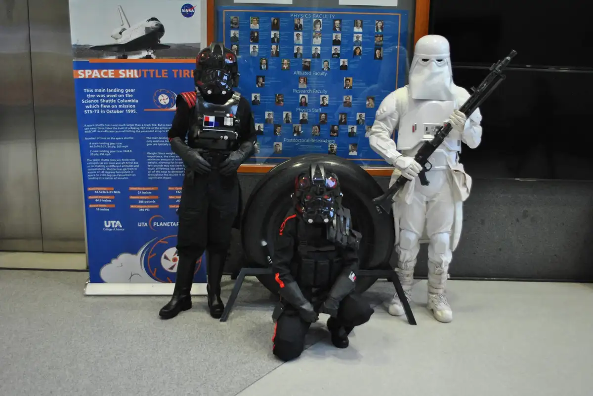 Three people in Star Wars costumes—a black-armored figure, a kneeling black-armored figure, and a white-armored figure…