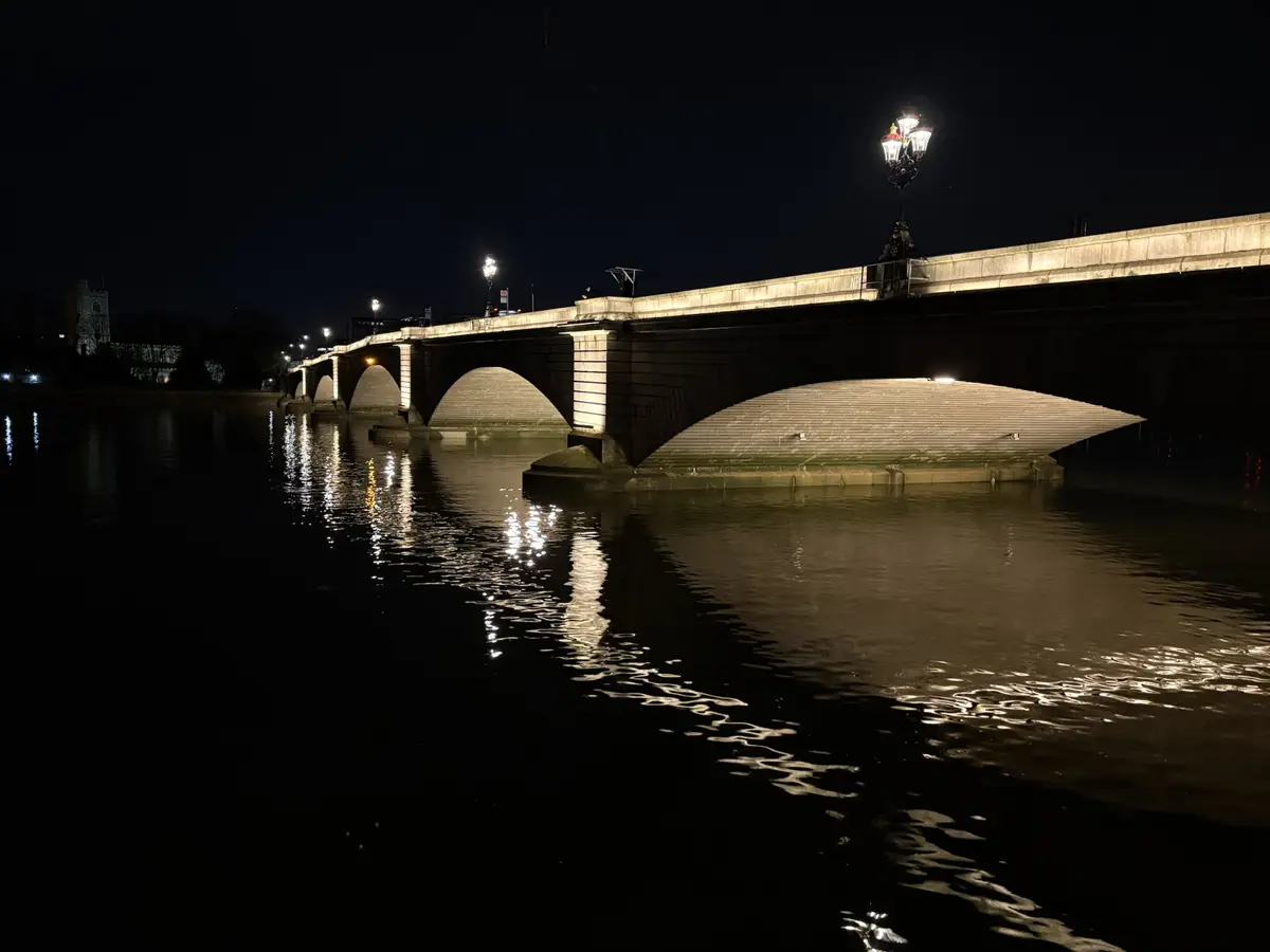 A nighttime photograph of a stone arch bridge over water, with streetlights illuminating its structure and casting…