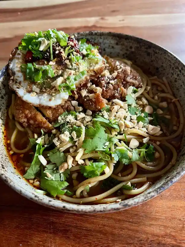 A close-up photo of a bowl of Asian-style noodles in a dark broth, topped with fried chicken, a sunny-side-up egg, chopped…