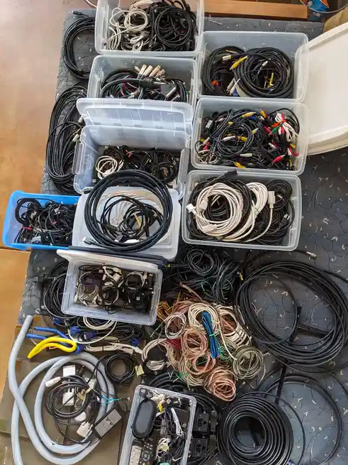 An overhead shot shows a large collection of tangled and neatly coiled electronic cables spread across a table, filling…