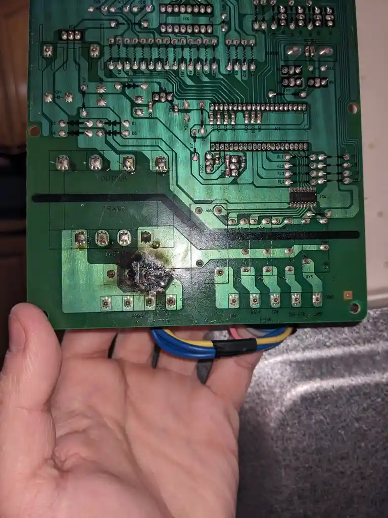 A hand holds a green circuit board with visible soldering and component labels like 'INPUT', 'OUTPUT', and 'TRANS'. A large…