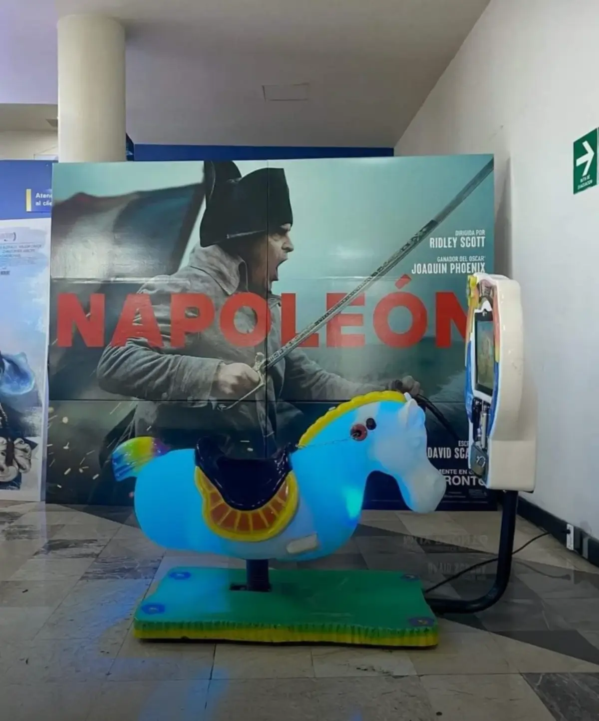 A bright blue, plastic children's ride-on horse stands in front of a large movie poster for 'Napoleon,' featuring actor…