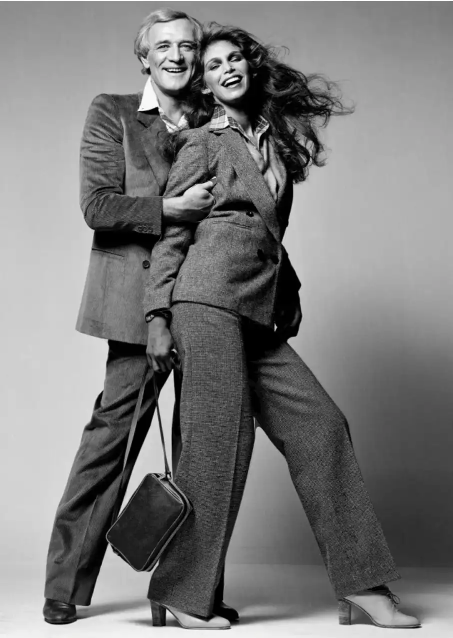 A black-and-white photo shows a smiling man in a suit embracing a woman in a matching suit, who is laughing with her hair…