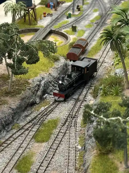 A detailed model of a black steam locomotive pulling two brown coal cars on a miniature railway track, surrounded by model…