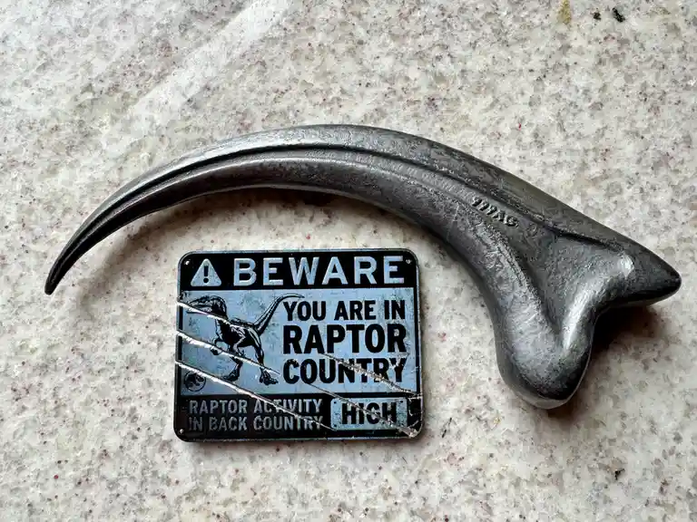 A large, curved, metallic raptor claw is placed over a warning sign that reads 'BEWARE YOU ARE IN RAPTOR COUNTRY' with…
