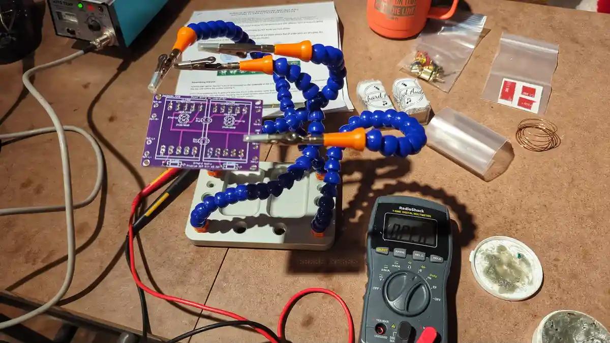 A close-up photo of an electronics workbench shows a purple circuit board mounted on a white stand, with blue flexible…