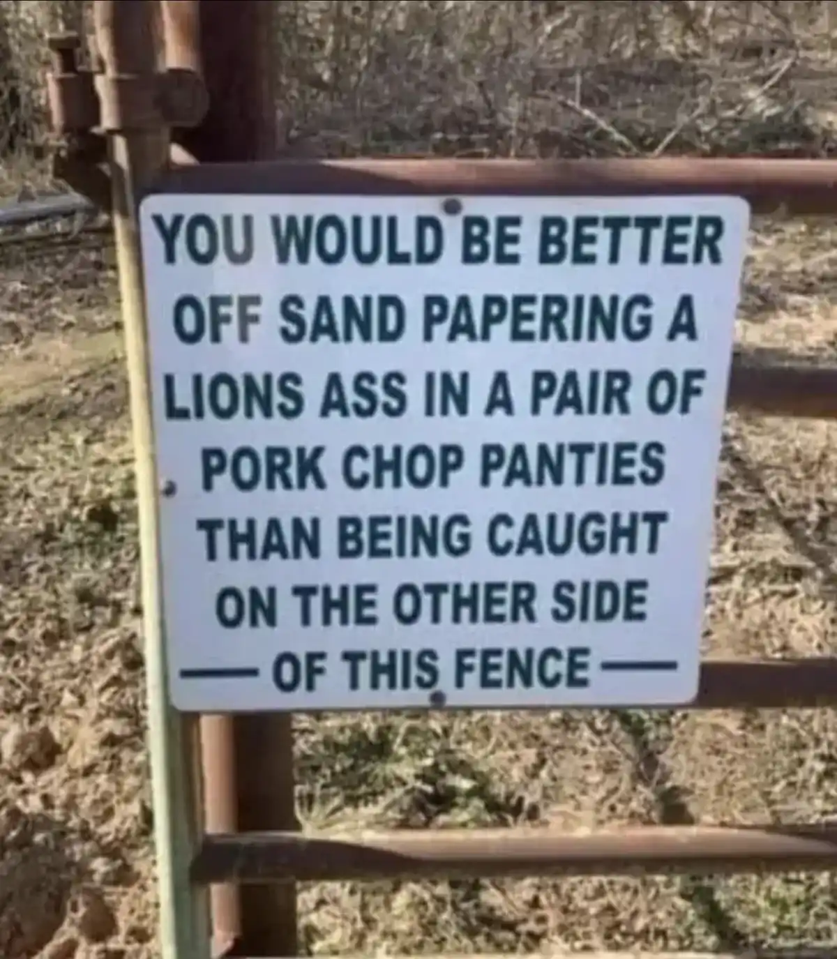 A white rectangular sign with bold black text is mounted on a rusty metal fence in a dry, grassy outdoor setting. The sign…