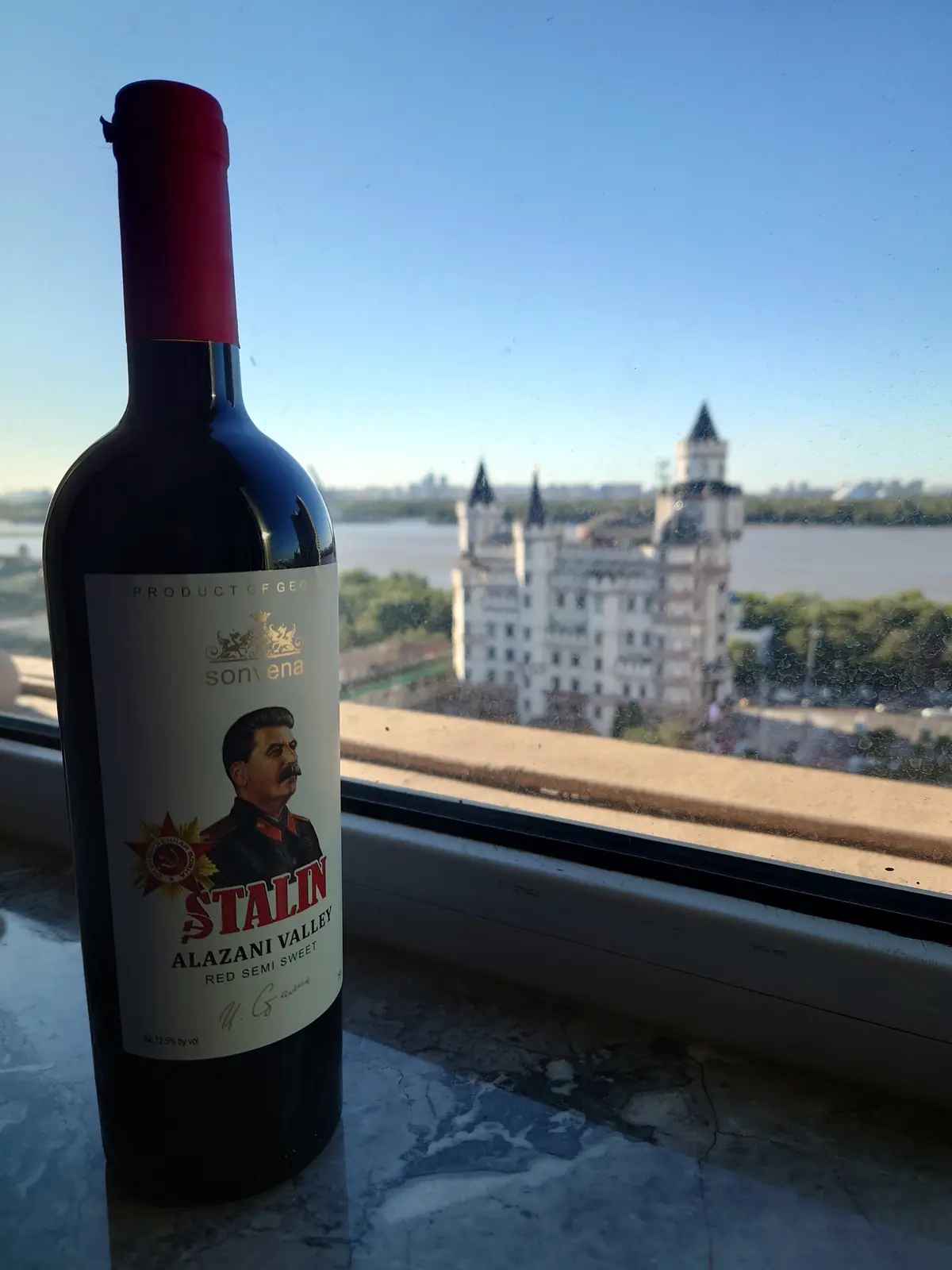 A bottle of 'Sonvena' wine labeled 'STALIN ALAZANI VALLEY RED SEMI SWEET' sits on a windowsill, with a blurry view of a…