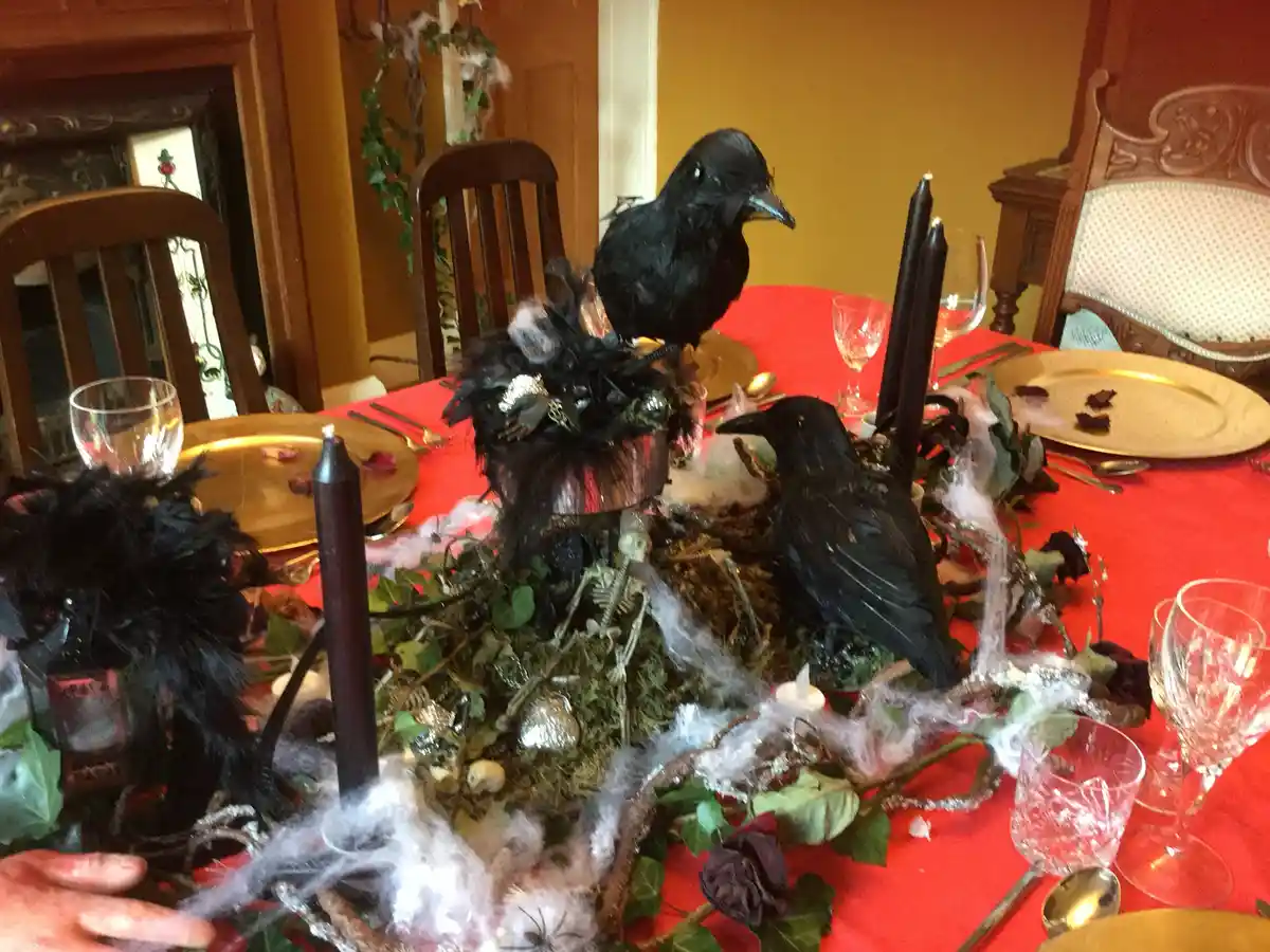 A dining table is set for a Halloween party with a red tablecloth, gold plates, and black candles. Two black crow figurines…
