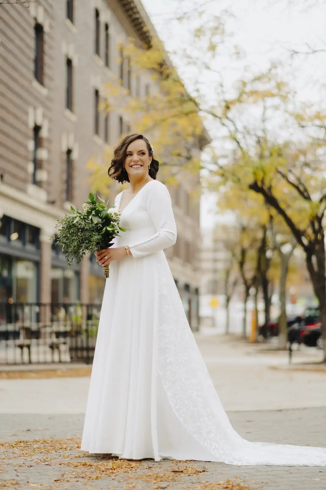 A smiling woman in a long white wedding dress stands on a city sidewalk, holding a green and white bouquet, with autumn…