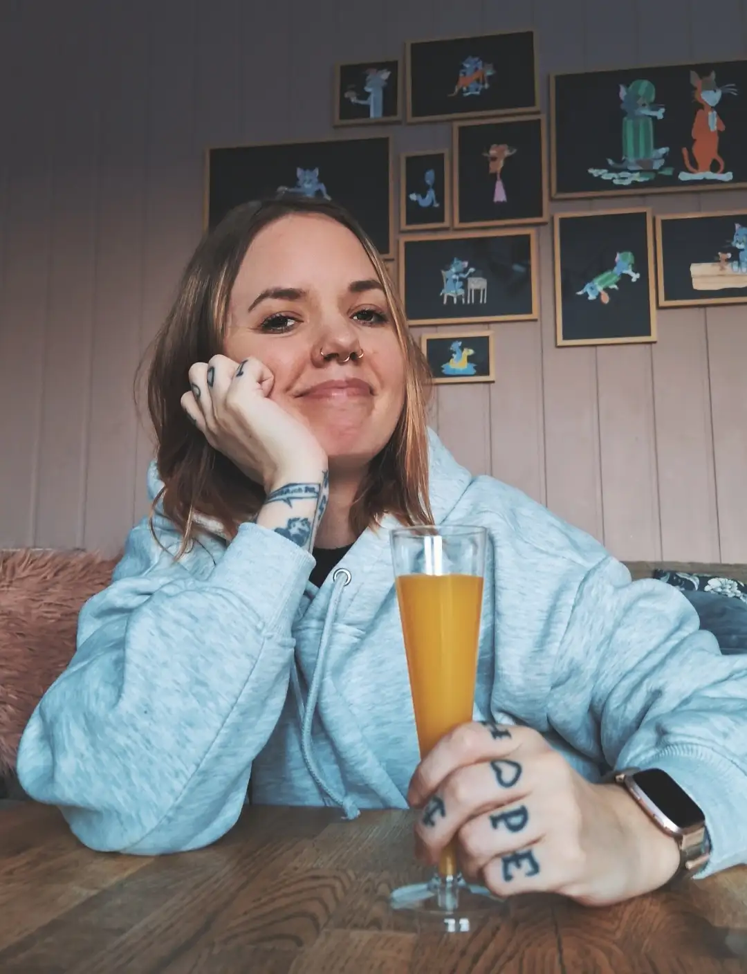 A woman with tattoos on her fingers spelling 'HOPE' and on her wrist sits at a wooden table holding a glass of orange…