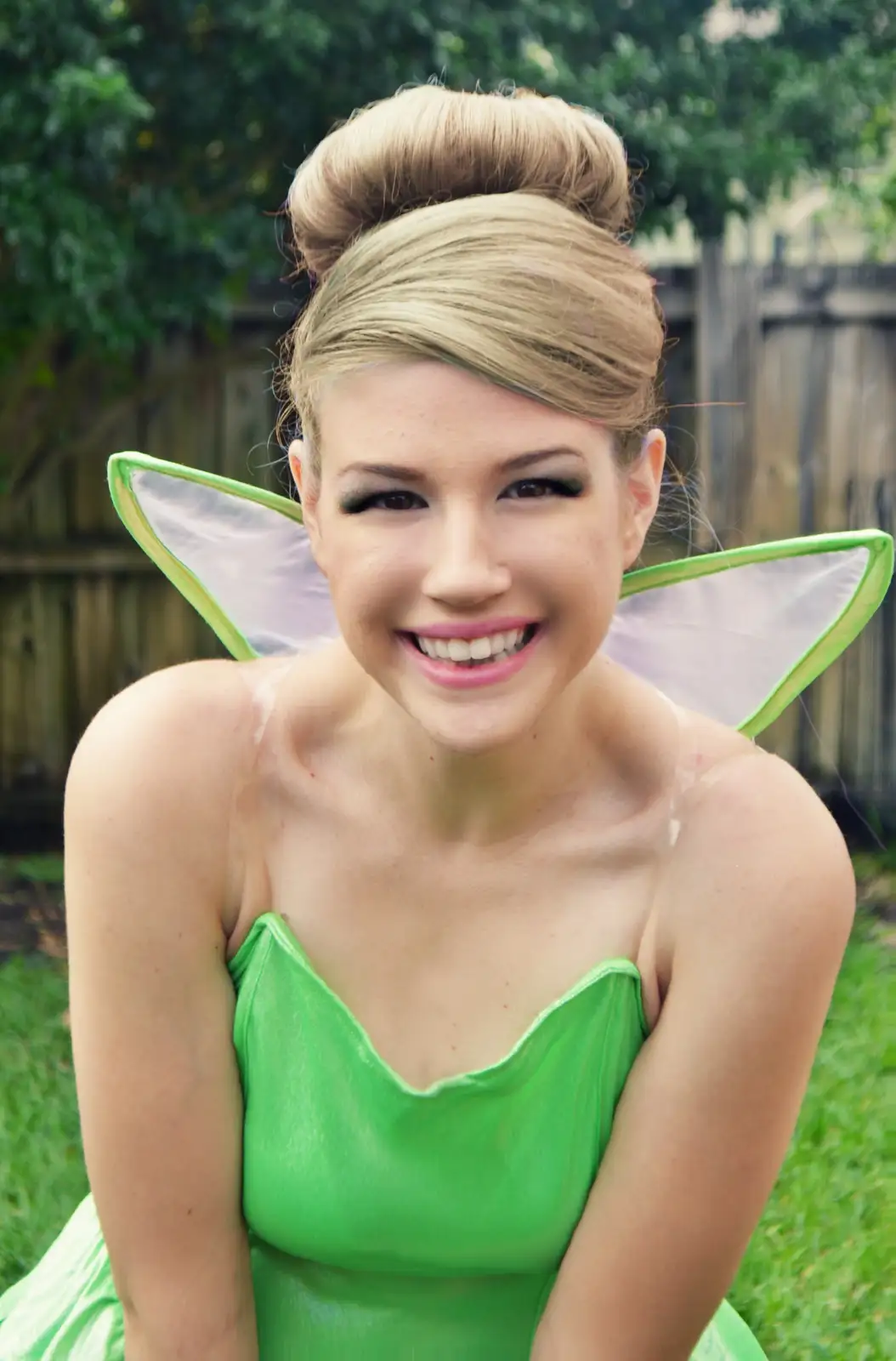A young woman with blonde hair in a high bun smiles at the camera while wearing a green fairy costume with transparent wings…