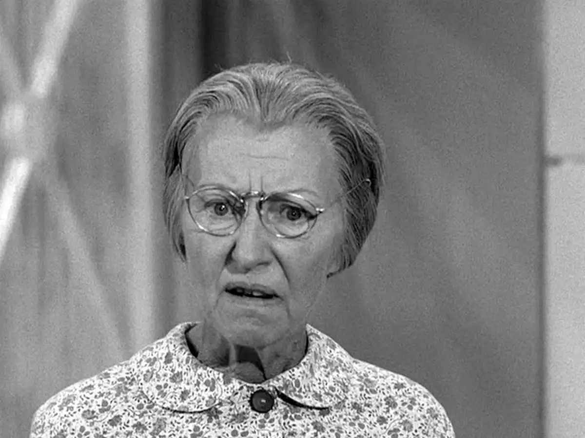 A black-and-white image of an elderly woman with glasses, wearing a patterned blouse, looking shocked or horrified. The…