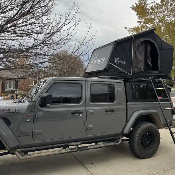A gray Jeep Gladiator pickup truck is parked on a residential driveway with a black rooftop tent installed and fully…
