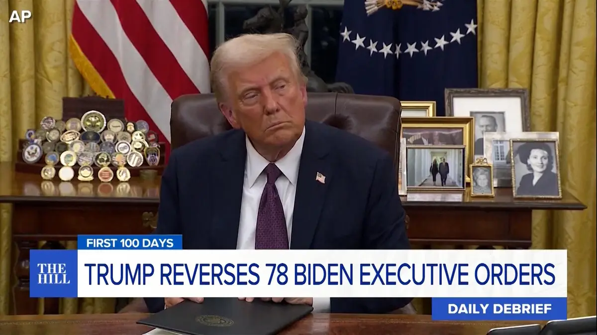 A still image of Donald Trump sitting at a desk in the Oval Office with a weary, unimpressed facial expression. Behind him…
