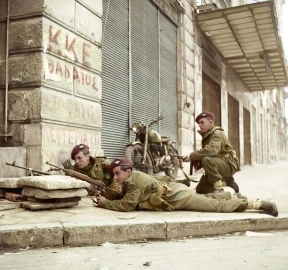 Three soldiers in olive uniforms and red berets are positioned on a sidewalk in a war-torn urban setting, with one prone…