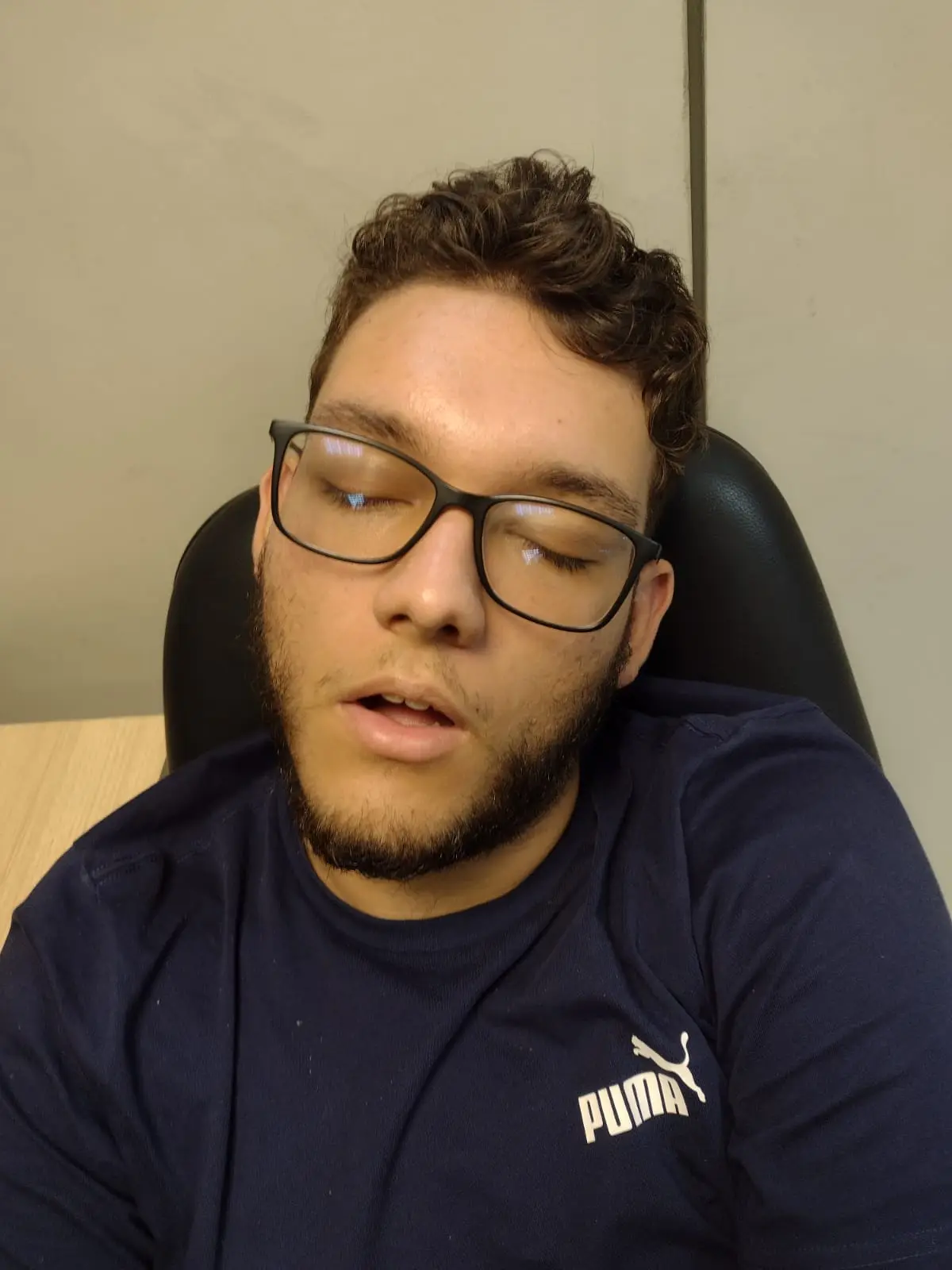 A man with dark curly hair, glasses, and a beard is slumped in a black office chair with his eyes closed and mouth slightly…