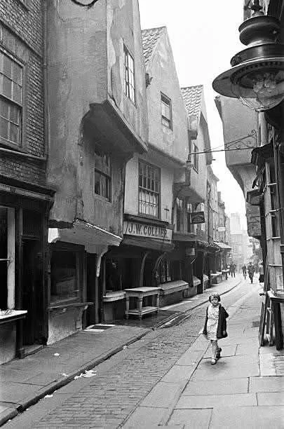 A black-and-white photo shows a young girl walking down a narrow, cobblestoned street lined with old, weathered buildings…