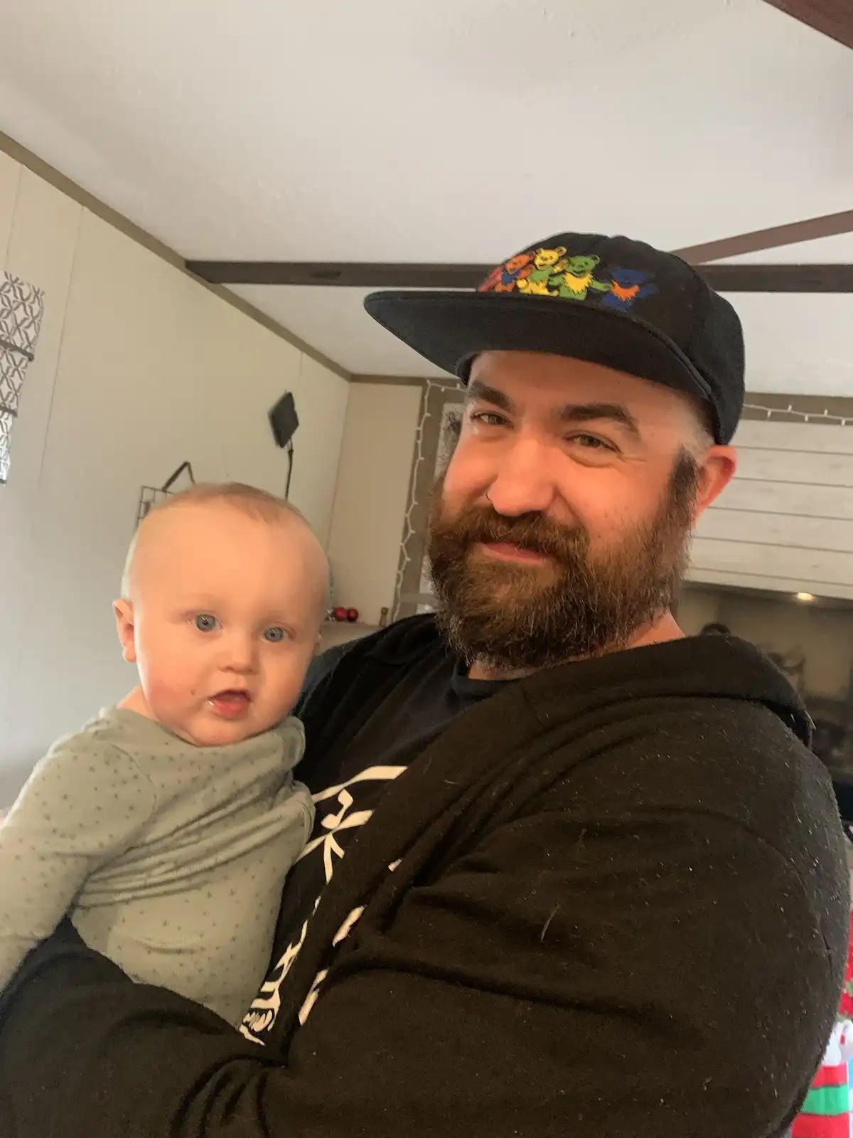 A bearded man wearing a black cap with colorful bear graphics holds a baby in a green onesie. The man has a smug…