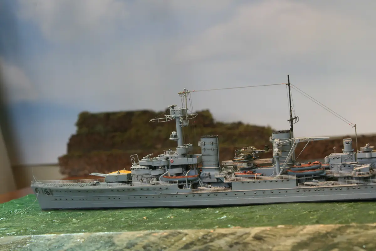 A highly detailed grey model of a warship, likely a German cruiser, sits on a green textured surface resembling water, with…