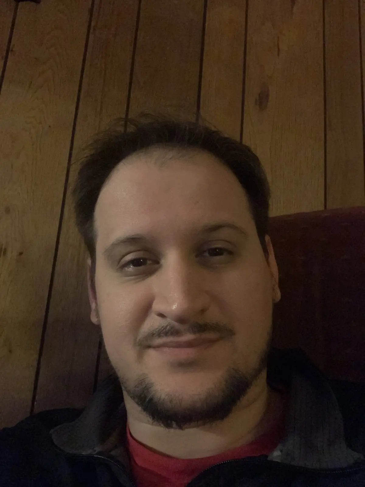 A close-up selfie of a man with dark hair, a goatee, and a slight smile, wearing a red shirt under a dark jacket, seated…