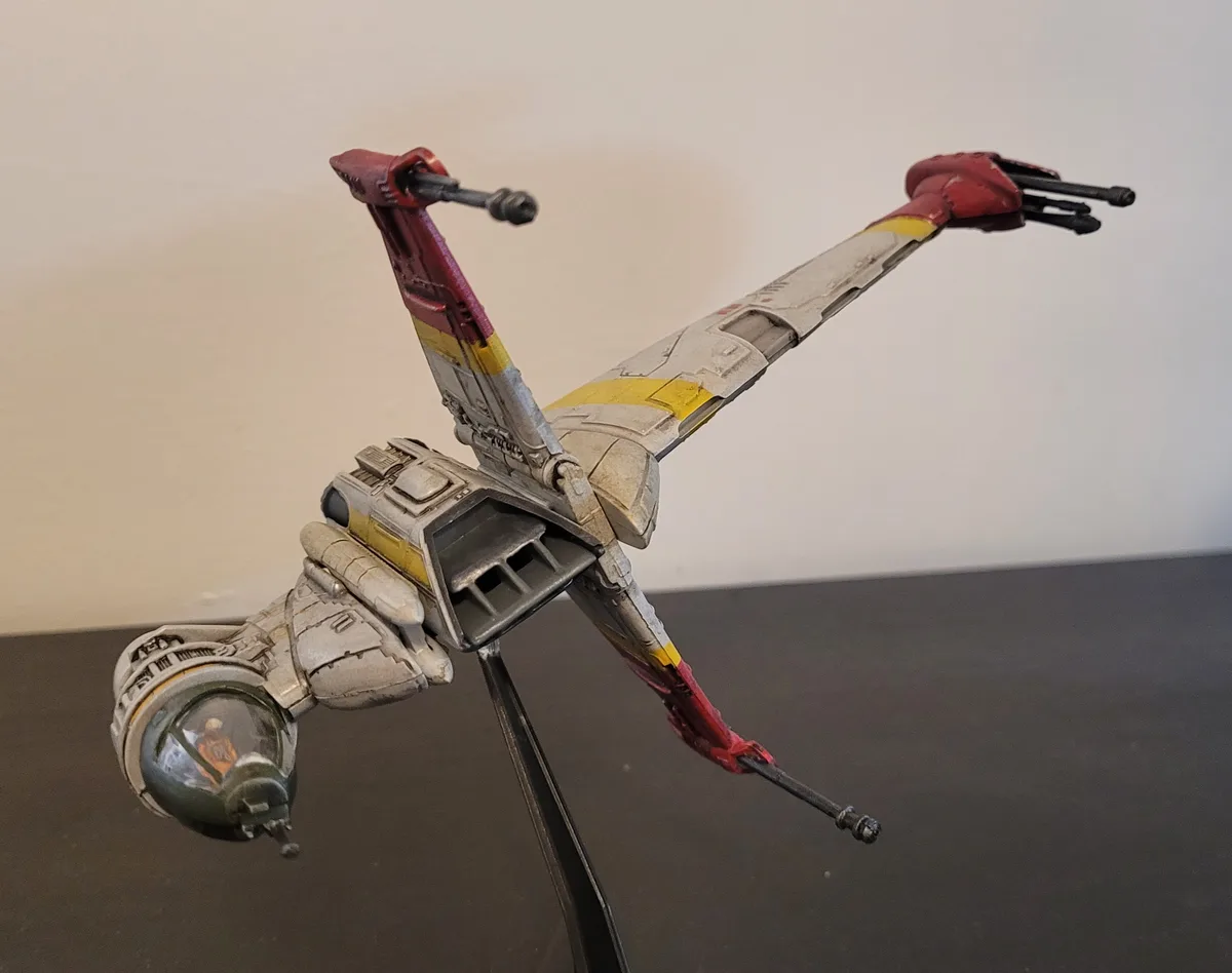 A detailed model of a Star Wars X-wing starfighter, painted with red and yellow markings and showing weathering, is…