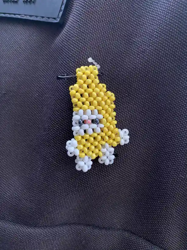 A handmade beadwork pin shaped like a yellow banana with a white face, black eyes, and a red nose, pinned onto a dark gray…
