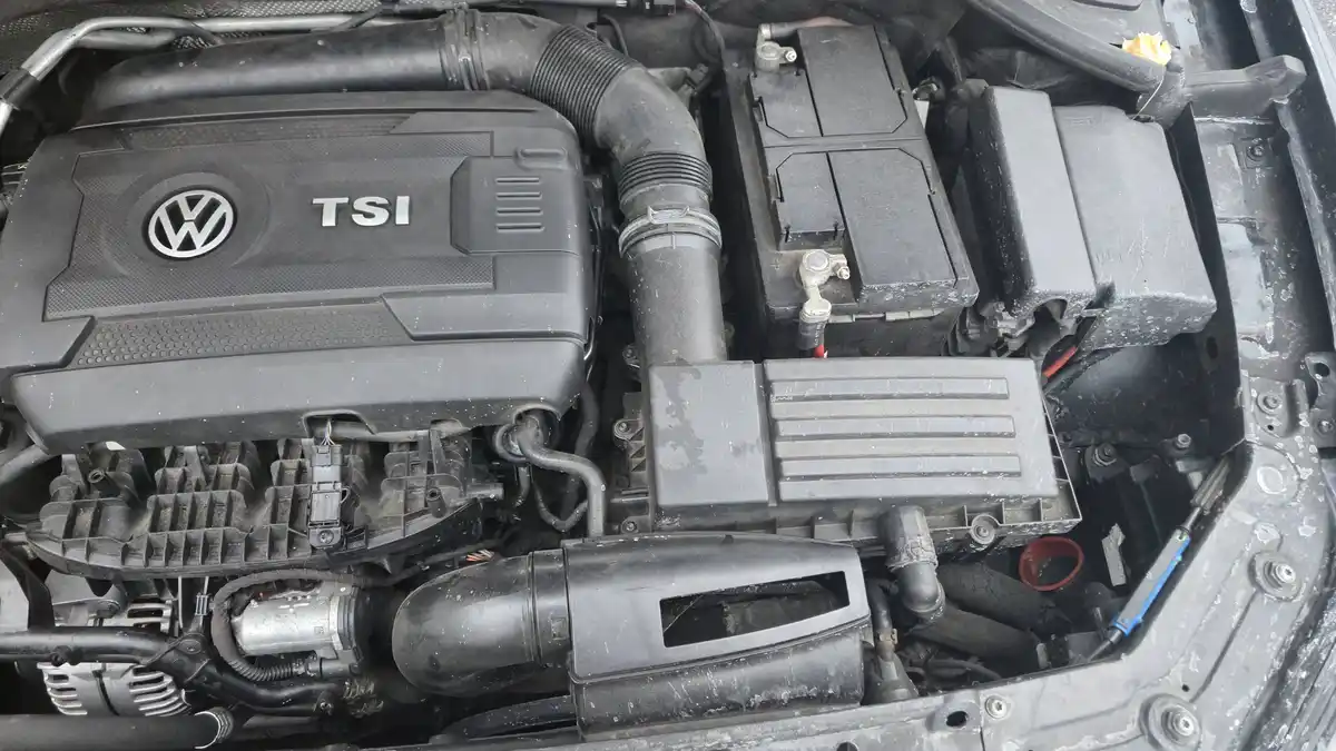 A close-up photo of a dirty Volkswagen car engine bay, showing the black engine cover with the VW logo and "TSI" text. The…