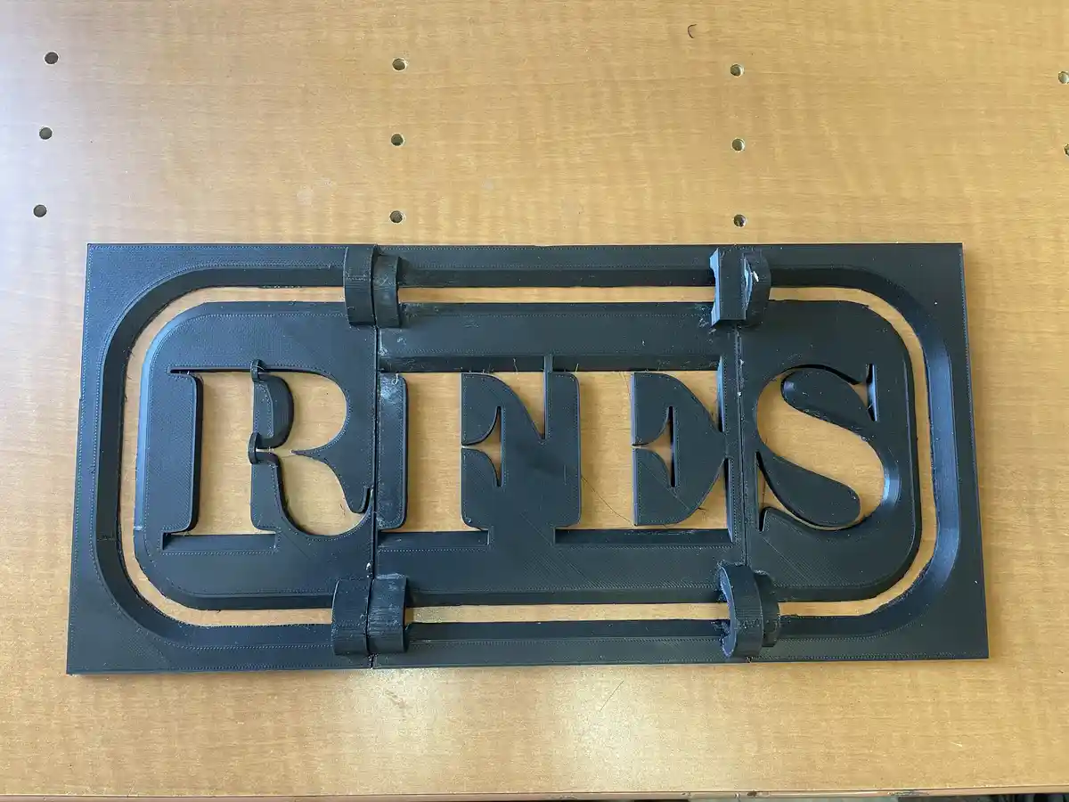 A black 3D-printed sign with the word 'DRINKS' in large, stylized letters sits on a light brown wooden table with visible…