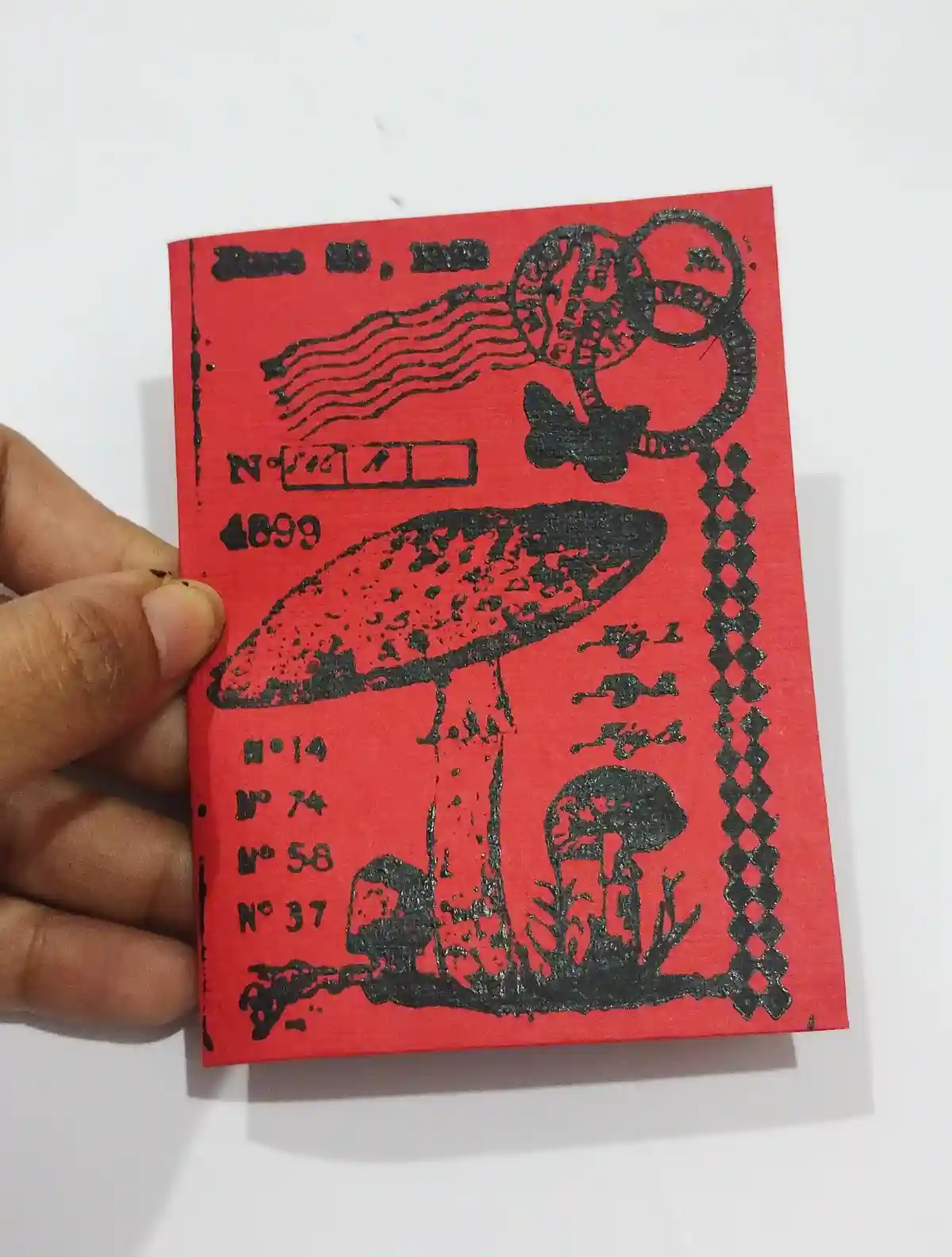 A hand holds a red, vintage-style postcard with black ink illustrations of mushrooms, a stylized globe, and decorative…
